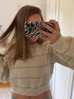 Beige stickad tröja med blå ränder - Mysig beige stickad tröja med tunna ljusblå ränder. Tröjan har rund hals, långa ärmar och ribbade muddar vid ärmslut och nederkant. Perfekt för lager på lager och chill stil.