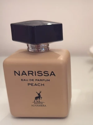 Narissa Peach Eau de Parfum - Narissa Peach Eau de Parfum från Maison Alhambra i en beige, fyrkantig flaska med svart, kantigt lock. Flaskan har stilren text och en elegant hjortlogga på framsidan. Doften har inslag av persika och passar dig som gillar fruktiga parfymer.