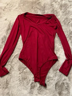Röd långärmad body med v-ringning - Snygg röd body med långa ärmar och v-ringad hals. Tillverkad i ett stretchigt och mjukt material som sitter skönt på kroppen. Perfekt att styla med jeans eller kjol för en trendig look.