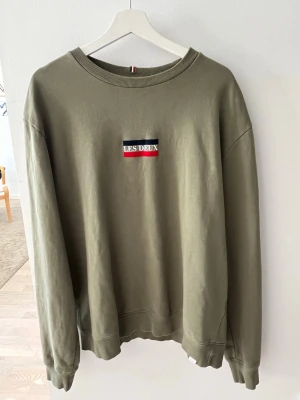 Olivgrön sweatshirt från Les Deux - Olivgrön sweatshirt från Les Deux med broderad logga i vitt, rött och marinblått på bröstet. Tröjan har rund halsringning, ribbade muddar vid ärmslut och nederkant samt en avslappnad passform. Perfekt för en casual look. Storlek XXL