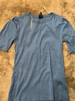 Blå ribbad topp med puffärm - Säljer en ljusblå ribbad topp med puffärm och rund hals. Toppen har halvlånga ärmar och är gjord i ett mjukt material som sitter bekvämt. Perfekt att styla med jeans eller kjol för en enkel men snygg look.