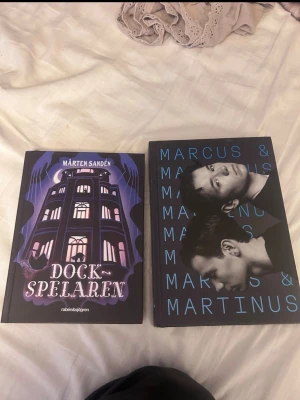 Dockspelaren & Marcus & Martinus bok - Säljer två böcker: 'Dockspelaren' av Mårten Sandén med ett mystiskt omslag i lila, svart och vitt samt en bok om Marcus & Martinus med svart omslag och blå text. Perfekt för dig som gillar spännande berättelser och musikbiografier.