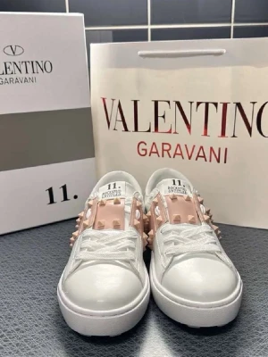 Valentino Garavani vita sneakers med nitar - Snygga vita sneakers från Valentino Garavani med ljusrosa band och ikoniska nitar på sidorna och hälen. Skorna har en chunky sula och är tillverkade i läder med vita skosnören. Perfekta för dig som vill sticka ut med en lyxig och trendig look.