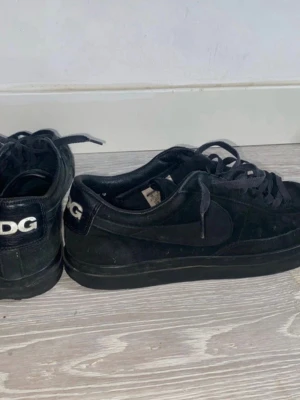 Svarta CDG x Nike sneakers - Säljer ett par svarta sneakers från Nike i samarbete med CDG. Skorna har CDG-logga i vitt på hälen, är låga och har ovandel i mocka och läder. Klassisk Nike-swoosh på sidan och svarta skosnören. Perfekta för dig som gillar stilrena och exklusiva sneakers.