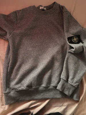 Grå sweatshirt från Stone Island - Säljer en klassisk grå sweatshirt från Stone Island med rund halsringning och ribbade muddar. Tröjan har den ikoniska Stone Island-loggan på vänster ärm. Perfekt för en avslappnad och stilren look.