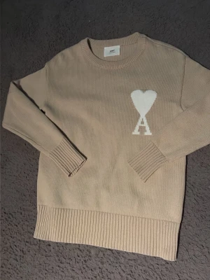 Beige stickad tröja med hjärtdetalj - Säljer en beige stickad tröja med rund hals och ribbade muddar. På bröstet finns en vit hjärtdetalj med bokstaven 'A' under. Tröjan har en avslappnad passform och är perfekt för lager-på-lager. Materialet känns mjukt och skönt mot huden.