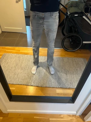Ljusgrå slim fit jeans dsquared2 - Säljer ett par ljusgrå slim fit jeans med slitningar och diskreta färgstänk. Jeansen har klassisk femficksdesign och knappgylf. Perfekta för en avslappnad och trendig look. Dsquared2 