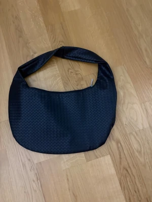 Svart flätad handväska - Säljer en svart handväska med flätad struktur och bred axelrem. Väskan har en mjuk, rundad form och är tillverkad i syntetmaterial. Perfekt för dig som vill ha en stilren och trendig accessoar till vardagslooken. Har ingen prislapp, men är i nytt skick har aldrig använt. Kan diskutera priser