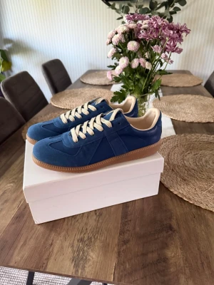 Blåa gats - Snygga blå sneakers med beige snörning och innersula. Skorna har en klassisk låg siluett och gummisula i brunt. Materialet är en mix av mocka och läder vilket ger en stilren och trendig look. Perfekta för dig som vill ha något unikt till din outfit.