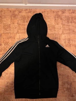 Svart Adidas Zip-Up - Tröjan är i storlek S men passar dessutom M, använt ett par gånger så den är i bra skick, perfekt för att använda till träning eller andra tillfällen. Skriv ifall ni har funderingar