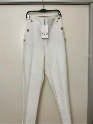Vita high waist jeans från Zara - Säljer ett par vita high waist kostymbyxor från Zara med raka ben och dekorativa guldknappar vid midjan. Snygg och modern passform som ger en clean look. Perfekta att matcha med crop tops eller oversized tröjor.