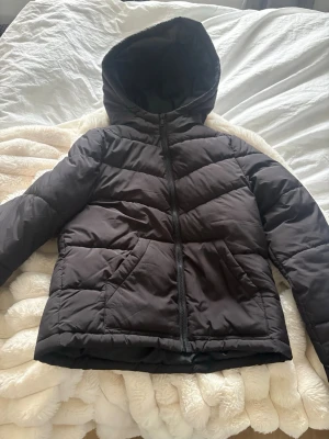 Svart pufferjacka med huva - Säljer en svart pufferjacka med dragkedja och stor huva. Jackan har två snedställda fickor framtill och quiltad design med breda paneler. Perfekt för kalla dagar och riktigt skön att bära. Materialet är syntet och jackan har en klassisk, rak passform.