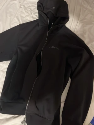 Svart hoodie från Sail Racing - Säljer en svart hoodie från Sail Racing med dragkedja framtill och huva. Hoodien har diskret logga på bröstet och är tillverkad i ett mjukt, stretchigt material som känns skönt mot huden. Perfekt för dig som gillar stilren och sportig look.