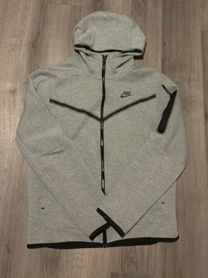 Ljusgrå Nike tech fleece hoodie med dragkedja - Snygg ljusgrå hoodie från Nike med hel dragkedja framtill och svarta detaljer. Har en huva, diskret Nike-logga på bröstet och en praktisk ficka med dragkedja på ärmen. Storlek M 