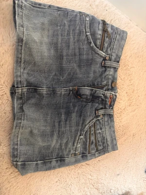 Mini jeanskjol med dragkedjor - Mini jeanskjol i blå tvättad denim med flera dragkedjedetaljer och orangea kontrastsömmar. Klassisk femficksmodell med bälteshällor och knappstängning. Perfekt för dig som gillar en avslappnad och trendig stil.