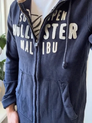 Hollister hoodie full zip M Mörkblå  - Snygg mörkblå hoodie från Hollister med dragkedja framtill och stora vita bokstäver på bröstet. Modellen har huva med dragsko och två fickor framtill. Perfekt för en avslappnad och cool stil. Mjuk bomullsblandning för extra komfort.