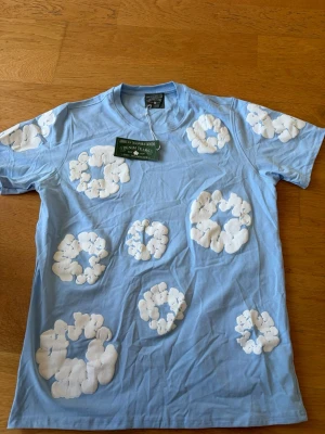 Blå t-shirt med vita blommor Denim Tears - Ljusblå t-shirt från Denim Tears med stora vita blommiga tryck över hela tröjan. Klassisk rund hals och korta ärmar. Tillverkad i mjuk bomull som känns skön mot huden. Perfekt för dig som vill sticka ut med en unik och trendig look.