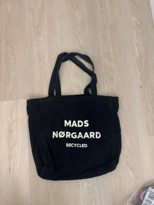 Svart shoppingväska Mads Nørgaard - Cool svart tygväska från Mads Nørgaard med trycket 'RECYCLED' på framsidan. Väskan har dubbla axelband och är tillverkad i återvunnet material. Perfekt för att bära med sig allt man behöver under dagen.