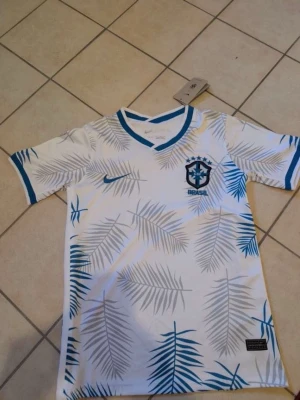 Vit Brasilien fotbollströja Nike - Snygg vit fotbollströja från Nike med Brasilien-emblem på bröstet. Tröjan har blå detaljer vid krage och ärmslut samt ett mönster av palmblad i grått och blått. Perfekt för dig som älskar fotboll och vill sticka ut på planen.