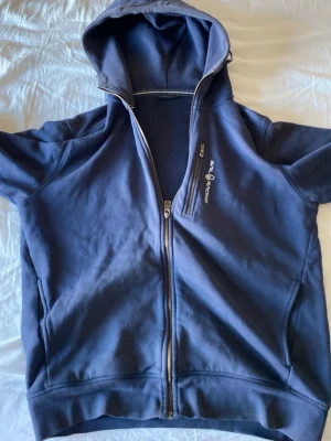 Marinblå hoodie från Sail Racing - Snygg marinblå hoodie från Sail Racing med dragkedja framtill och huva. Tröjan har två fickor på sidorna och diskret logga på bröstet. Det ända som är märkvärdigt är den lilla solblekningen på luvan annars perfekt
