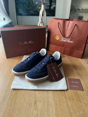 Loro Piana blå sneakers i mocka - Snygga sneakers från Loro Piana i mörkblå mocka med vita detaljer och ljusbrun sula. Klassisk låg modell med matchande blå skosnören och diskret design. Perfekta för dig som gillar exklusiva och stilrena skor.