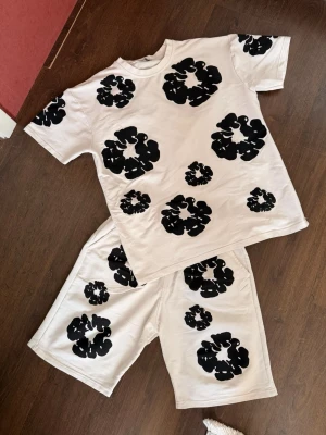 Vitt set med svarta blommor - Säljer ett vitt set med t-shirt och shorts, dekorerat med stora svarta blommönster över hela plaggen. T-shirten har rund hals och korta ärmar, shortsen har resår i midjan och fickor. Materialet ser ut att vara mjukt bomullstyg, perfekt för en avslappnad look.