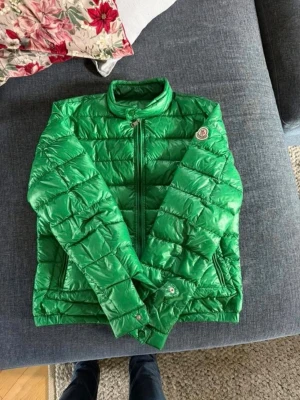 Moncler - Snygg grön pufferjacka från Moncler med glansigt material och quiltad design. Jackan har två framfickor med dragkedja, Moncler-logga på ärmen. En perfekt vår jacka. Storlek 2/ m
