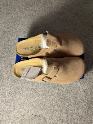 Beige Birkenstock tofflor i mocka - Säljer ett par snygga beige tofflor från Birkenstock i mjuk mocka. Modellen har en klassisk design med justerbart spänne och öppen häl. Perfekta för chill dagar hemma eller när du vill ha det bekvämt. Levereras i originalkartong.