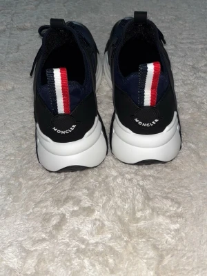 Moncler sneakers med blå detaljer - Snygga sneakers från Moncler i marinblått med svarta och vita detaljer samt en bred hälflik i rött, vitt och blått. Skorna har en chunky vit sula och ovandel i textil och syntet. Perfekta för dig som gillar sportig och modern stil.