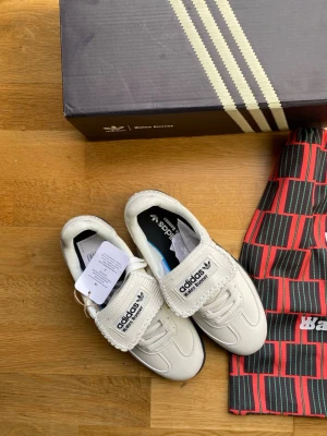 Adidas Wales Bonner vita sneakers - Unika vita sneakers från Adidas x Wales Bonner med svarta detaljer och guldfärgade nitar på sidan. Skorna har klassisk låg siluett, grovsydda detaljer och branding på plösen. Kommer med originalkartong och specialdesignad påse.