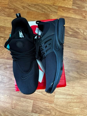 Svarta Nike Presto sneakers - Snygga helsvarta Nike Presto sneakers med mesh-ovandel och plastdetaljer på sidorna. Skorna har rund tå, klassisk snörning och diskret Nike-logga på tån och sidan. Perfekta för dig som gillar stilrena och sportiga sneakers.