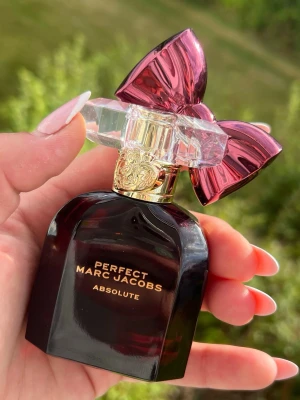 Perfect Marc Jacobs Absolut parfym - Säljer en elegant parfym från Marc Jacobs, Perfect Absolut. Flaskan är mörklila med gulddetaljer och har en unik kork med en stor rosa rosett i plast. Perfekt för dig som vill ha en lyxig och iögonfallande doftflaska i samlingen.