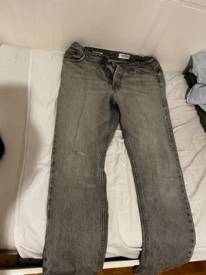 Jack & Jones W30L32 - Snygga grå jeans med rak passform och knäppning framtill. Jeansen har klassiska fem fickor och subtila slitningar på ena benet. Perfekta för en avslappnad och trendig look.