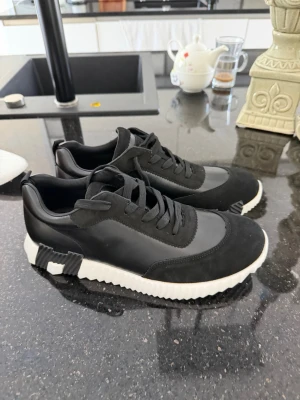 Svarta sneakers med vit sula - Stilrena svarta sneakers med vit, räfflad sula och svarta detaljer. Ovandelen är i en mix av mocka och slätt material, med svarta skosnören och låg profil. Sulan har en cool design med svarta H-liknande mönster undertill. Perfekta för en trendig vardagslook.
