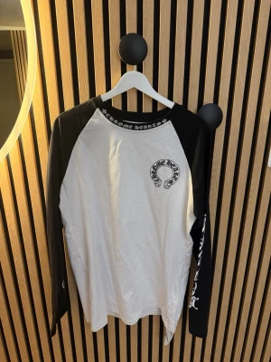 Svartvit långärmad topp Chrome Hearts - Säljer en svartvit långärmad topp från Chrome Hearts med tryck på bröstet och textdetaljer runt kragen. Ärmarna är svarta medan kroppen är vit, och materialet känns som mjuk bomull. Perfekt för dig som gillar streetwear och snygga detaljer.