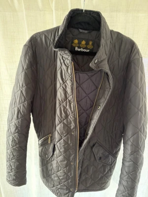 Svart quiltad jacka från Barbour - Stilren svart quiltad jacka från Barbour med dragkedja och tryckknappar framtill. Jackan har två fickor med lock och en snedställd ficka med dragkedja. Klassisk krage och rutmönstrad sömnad ger en tidlös look. Perfekt för kyligare dagar och höst. Storlek XS men skulle rekommendera S/M