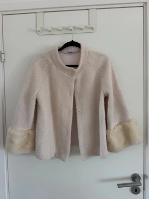 Ljusbeige kappa med fuskpälsdetaljer - Stilren ljusbeige kappa med tre stora knappar framtill och breda ärmar. Kappan har mjuka fuskpälsdetaljer vid ärmsluten som ger en lyxig känsla. Materialet är strukturerat och kappan har en avslappnad, något A-linjeformad passform. Använd förtal gånger, fortfarande i fint skick, endast en fläck på ena päls ärmen men inget som ska påverka hur den ser ut på, passar xs/s