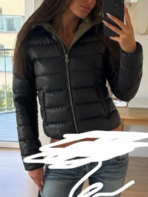 Svart pufferjacka med dragkedja - Snygg svart pufferjacka med quiltad design och dragkedja framtill. Jackan har hög krage och två sidofickor. Insidan är beige och materialet ser glansigt ut, vilket ger en modern look. Perfekt för kalla dagar och enkel att matcha med olika outfits.