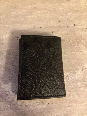Plånbok - Snygg svart plånbok med klassiskt monogrammönster i präglat läder. Kompakt och stilren design som passar perfekt i fickan. Perfekt accessoar för dig som gillar exklusiva detaljer och tidlös stil.