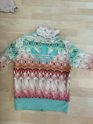 Färgglad Adidas ziptröja med fjärilar - Säljer en unik Adidas ziptröja med färgglatt fjärilsmönster i grönt, orange, rosa och beige. Tröjan har turkosa muddar, dragkedja framtill, två fickor och Adidas logga på ryggen. Perfekt för dig som vill sticka ut på träningen eller i vardagen.