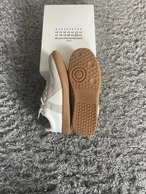 Maison Margiela vita sneakers - Snygga vita sneakers från Maison Margiela med beige detaljer och klassisk gummisula. Skorna har en låg silhuett, ovandel i läder och mocka samt vita skosnören. Perfekta för dig som gillar minimalistisk och stilren design.