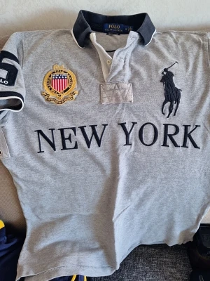 Grå pikétröja från Polo Ralph Lauren storlek M  - Säljer en grå pikéskjorta från Polo Ralph Lauren med stora broderade detaljer, svart logga och texten NEW YORK på bröstet. Skjortan har korta ärmar, krage och knappar framtill samt broderad patch på ärmen. Lite tecken på långvarig användning men påverkar absolut inte tröjans funktion.