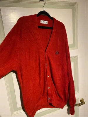 Röd kofta från IZOD - Röd kofta från IZOD med v-ringning och knäppning framtill. Klassisk design med broderad logga på bröstet och mjukt, tunt stickat material. Perfekt för lager på lager och enkel att matcha med olika outfits.