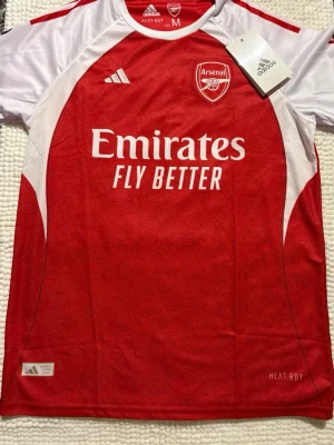 Arsenal matchtröja Adidas röd/vit - Säljer en officiell Arsenal matchtröja från Adidas i rött och vitt med klubbmärke och Emirates Fly Better-tryck på bröstet. Tröjan har korta ärmar, rund hals och är tillverkad i ett lätt, tekniskt material med HEAT.RDY-teknologi för bästa komfort. Köpt utomlands 