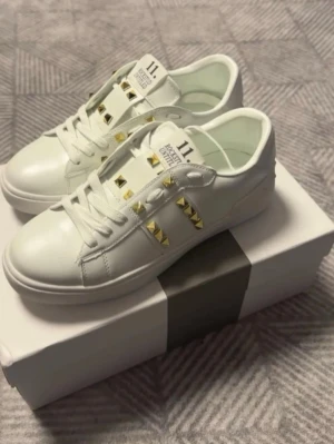 Vita sneakers med gulddetaljer - Säljer ett par vita sneakers med coola guldfärgade nitar på sidorna och över snörningen. Skorna har en chunky sula och klassisk låg profil. Tillverkade i slätt läderliknande material och har vita skosnören. Perfekta för dig som vill sticka ut med detaljer!