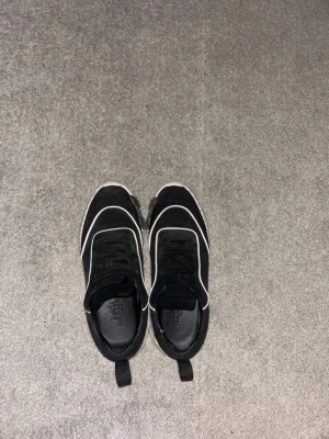 Svarta sneakers med vit sula - Svarta sneakers med vita detaljer och tjock vit sula. Skorna har en sportig look med svarta skosnören och en praktisk ögla bak på hälen. Perfekta för en avslappnad streetstyle.