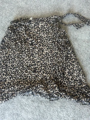 Leopardmönstrad omlottkjol från Lindex - Säljer en leopardmönstrad kort omlottkjol från Lindex med volangdetaljer och knytband i midjan. Kjolen är i beige och svart och har en riktigt trendig vibe. Perfekt för dig som vill sticka ut lite extra.