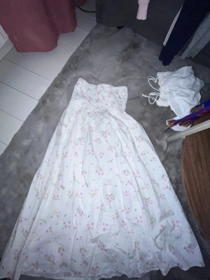 Vit blommig långklänning Gina Tricot - Supersöt vit långklänning från Gina Tricot med blommigt mönster i rosa och grönt. Klänningen är axelbandslös och har smock upptill för en bekväm passform. Perfekt för sommarens alla tillfällen!