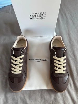 Maison Margiela bruna sneakers - Säljer ett par bruna sneakers från Maison Margiela med beige snörning och klassisk gummisula. Skorna har en stilren design med inslag av både mocka och skinn, vilket ger en lyxig känsla. Perfekta för dig som gillar minimalistisk och trendig stil.