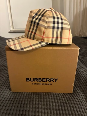 Burberry keps - Säljer en ikonisk Burberry keps i beige med det klassiska rutmönstret i svart, vitt och rött. Kepsen har böjd skärm och justerbar snapback baktill. Märkeslapp med 'Burberry London England' på baksidan. Perfekt accessoar för en trendig streetwear-look.